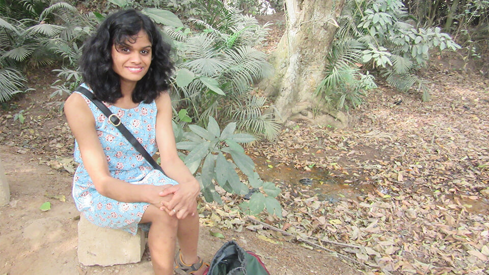 Female PhD student in a dress sat outside with greenery all around, smiling at the camera.