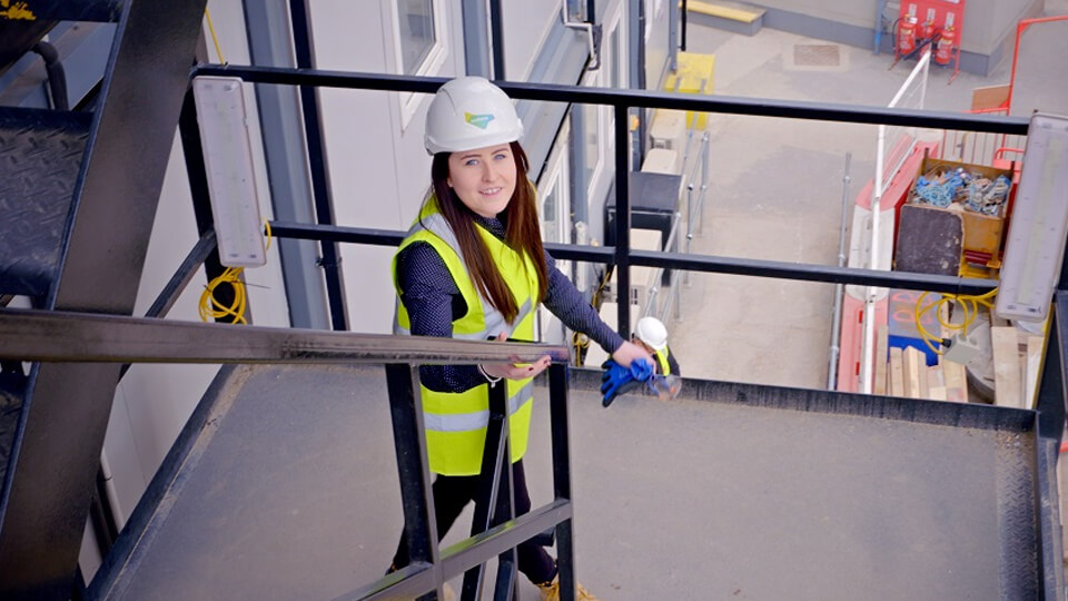 Female student in a high vis jacket and a white helmet standing on the stairs looking up at the camera with a building site in the background.