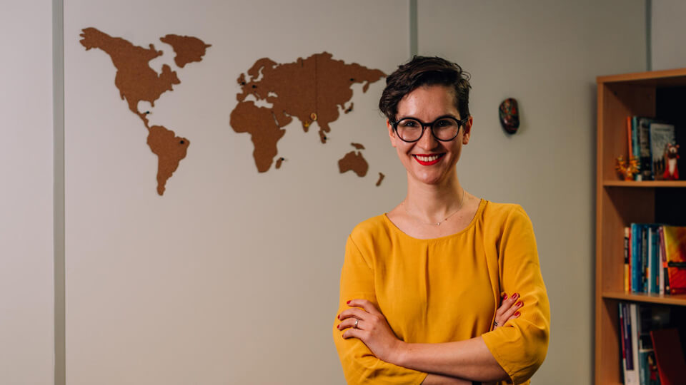 Female academic smiling at the camera, wearing a yellow top, with a world map in the background.