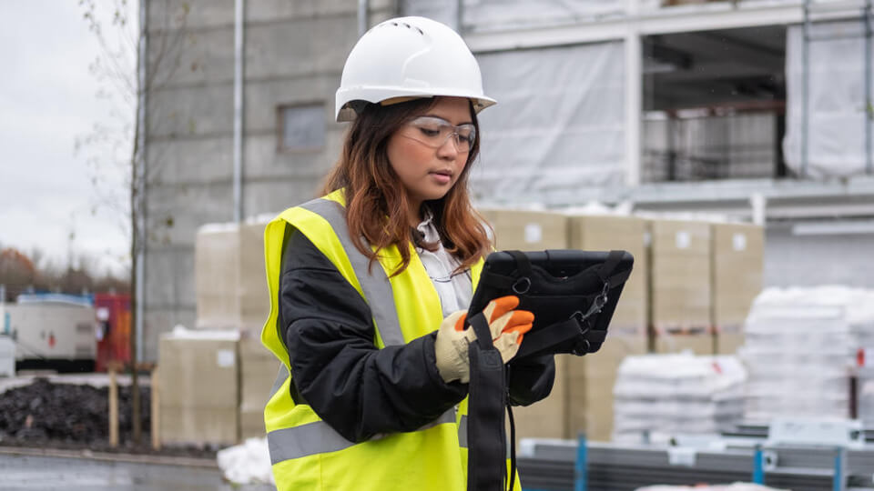 Female student at a site with a protective helmet and Hi-vis vest, looking at a tablet.