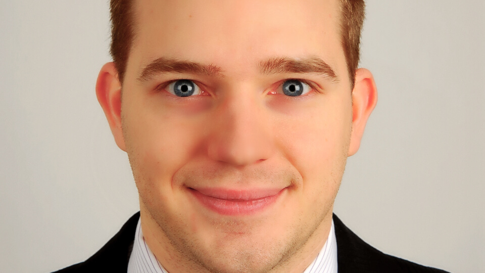 Male student in a suit standing in front of a white wall and smiling at the camera.