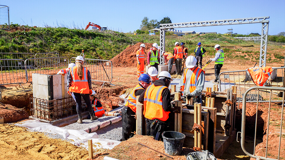 Group of students and staff on-site wearing high vis vests and white helmets, building a road on a sunny day.