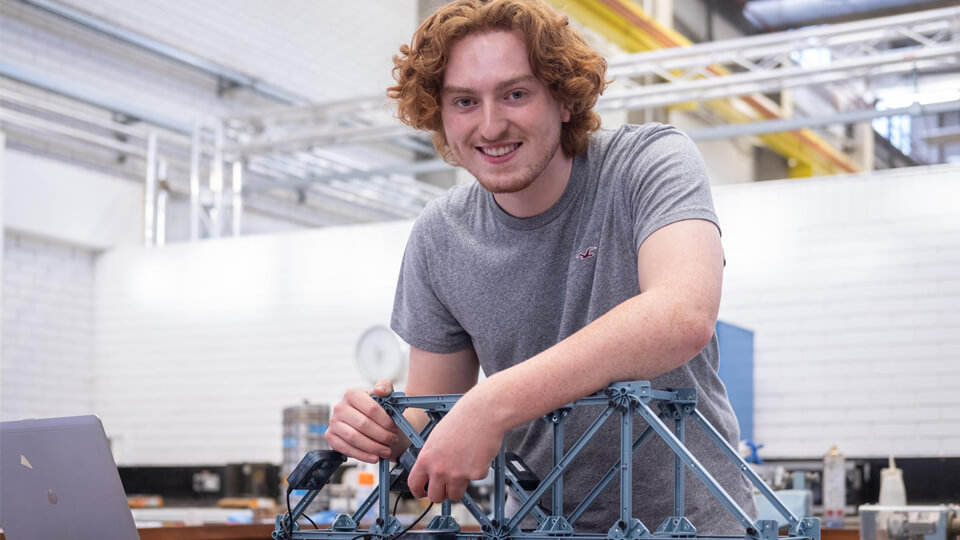 Male student smiling at the camera while working on a small-scale bridge model in a laboratory.