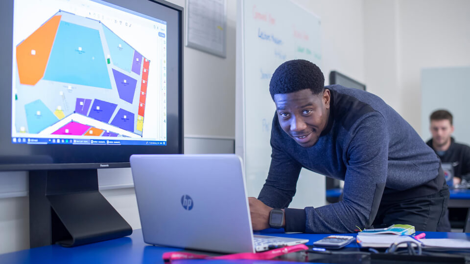 Male student smiling at the camera while working on his laptop in a classroom, with a screen with his project in the background.