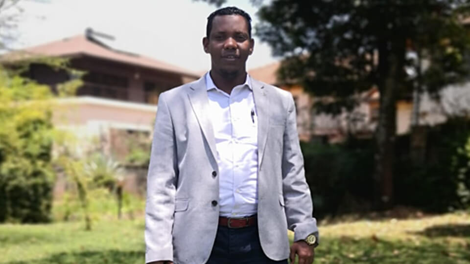 Male student standing outside in the sunshine in a suit with greenery all around him.