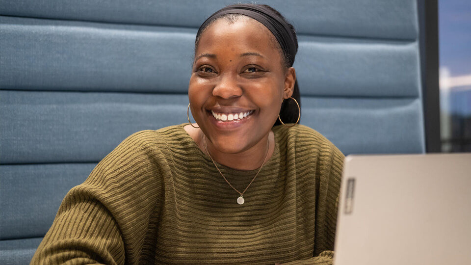 Female student sat on a couch in the STEMLab with her laptop, smiling at the camera, wearing a green jumper.