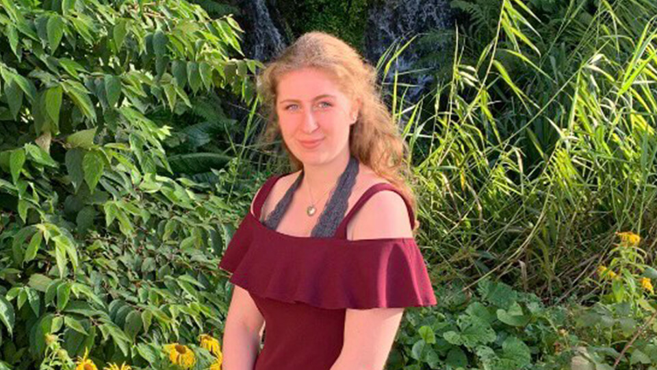 Female student standing in front of greenery, smiling at the camera outdoors.