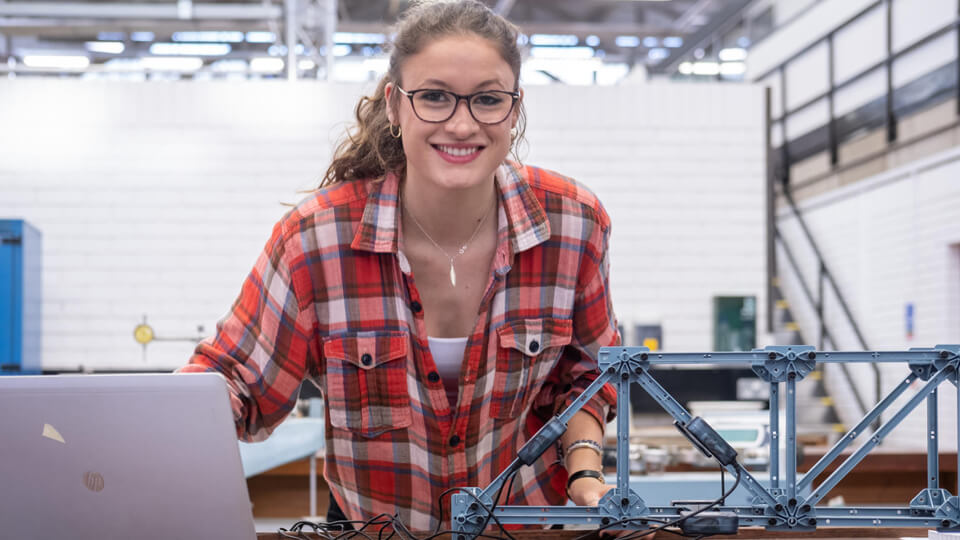 Female student in a built environment laboratory, smiling at the camera, wearing glasses and a red top.