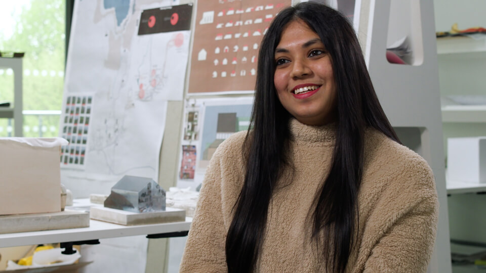 Female student sitting in a studio, smiling while talking to someone.