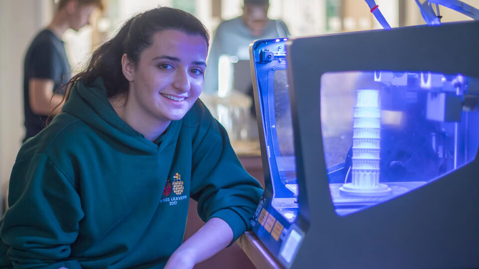 Female student in a 3D printing lab smiling at the camera next to a 3D printer that has finished printing a small model of the Tower of Pisa.