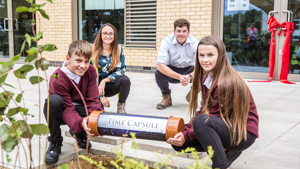 Student Kelly and her coworker squatting outside with two students, holding a time capsule in front of a School she helped build.
