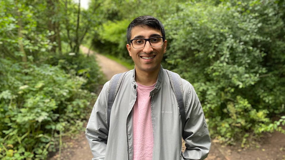 Male student in a forest smiling at the camera, wearing a backpack and glasses.