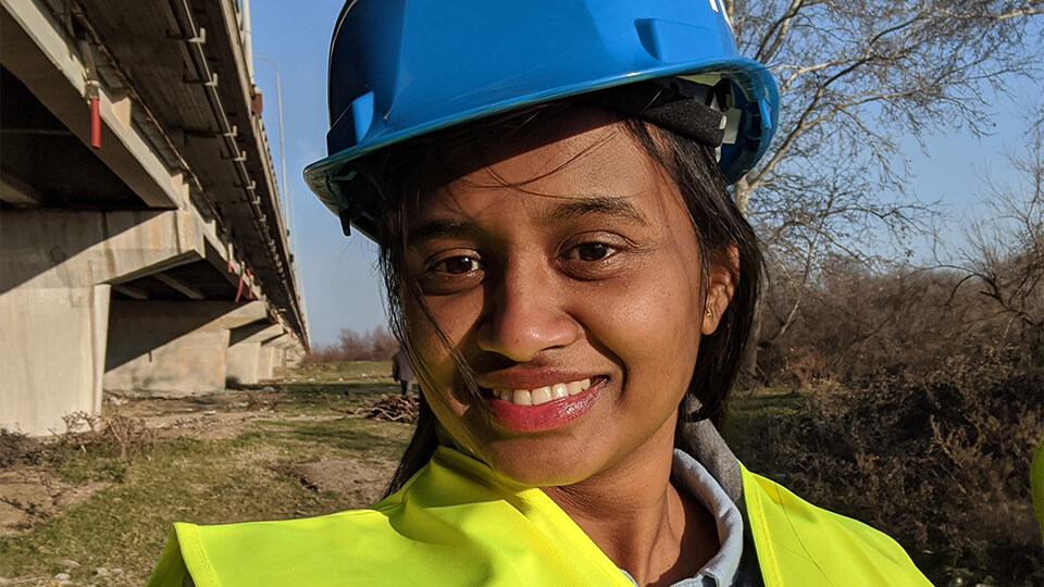 Female PhD student outside in a yellow high vis jacket and a blue safety helmet, smiling at the camera.