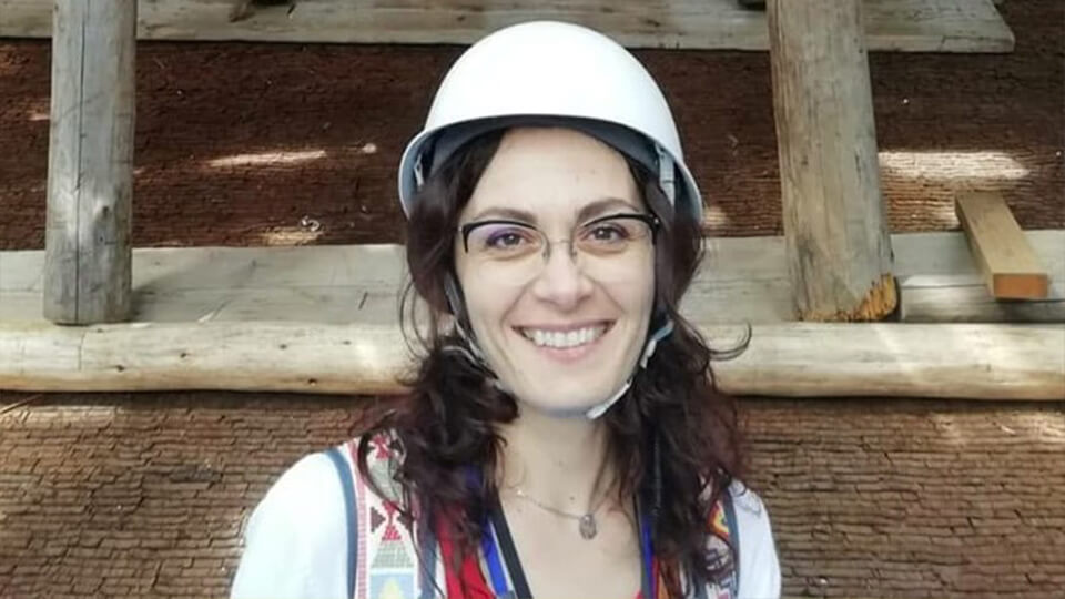 Female PhD student stood outside with a white protective helmet, smiling at the camera, with wooden beams in the background.