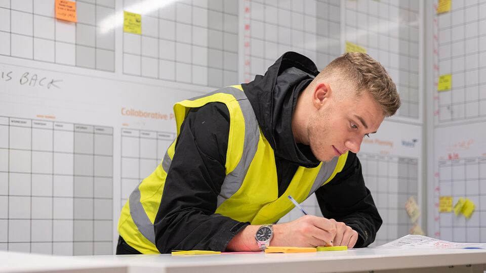 Male student in a high vis jacket in an office, writing down on commercial management plans.