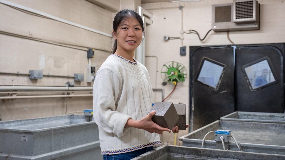 Female student in the concrete laboratory next to large water baths, holding a concrete cube.