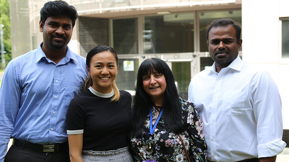 Three students and a member of staff standing in front of a building outside on a sunny day, smiling at the camera.