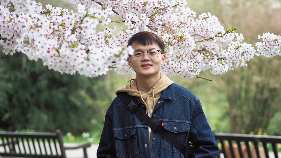 Male PhD student in a blue denim jacket, wearing glasses, standing outside in nature, smiling at the camera.