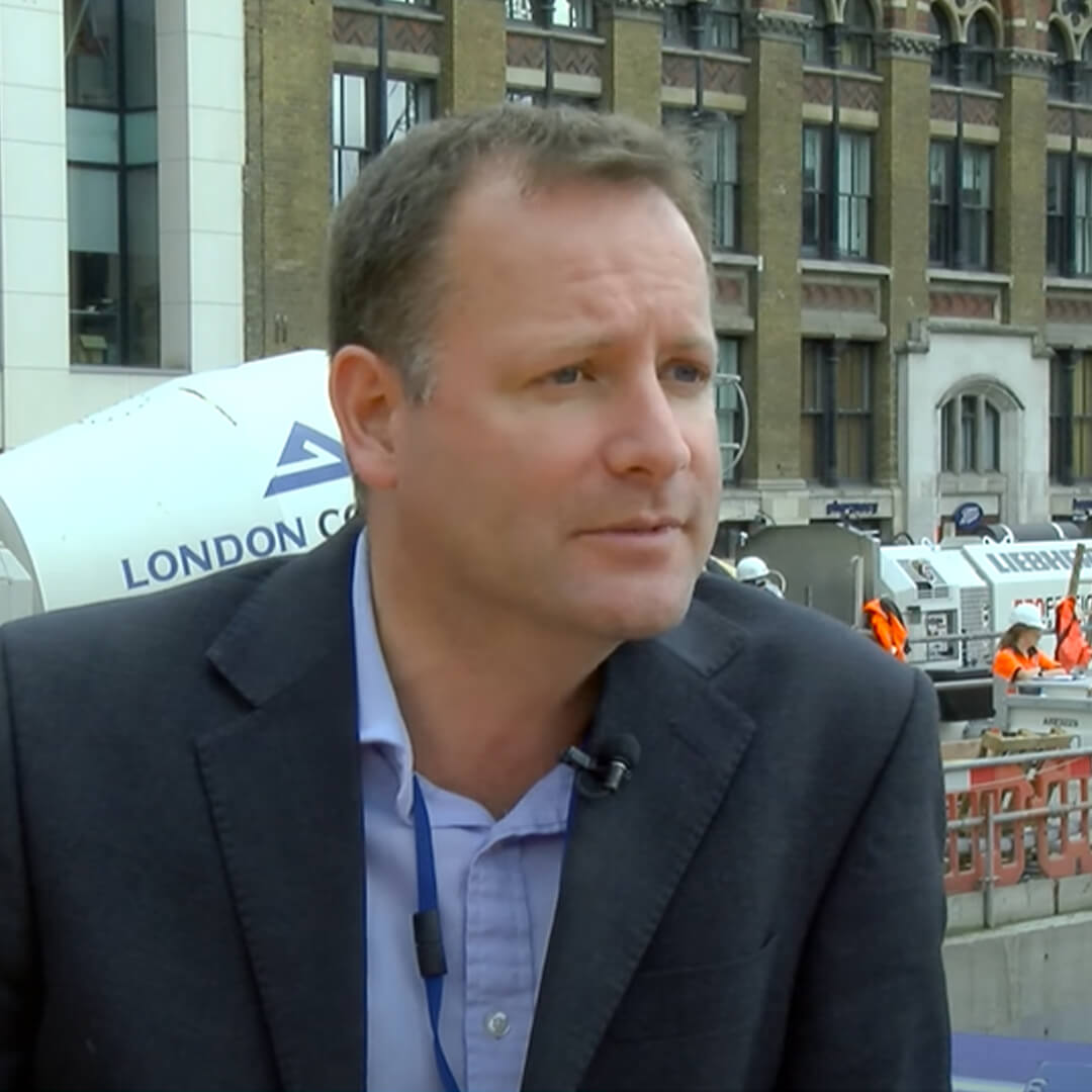 Man outside speaking to the camera with a building site in the background, wearing a suit and a lanyard.