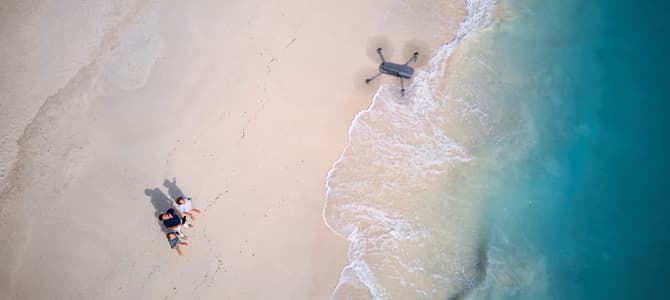 Drone looking over a sandy beach on a sunny day with clear water with three people looking up at it.