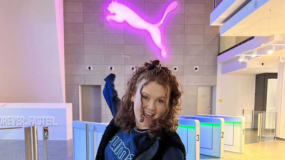 Student on placement at Puma standing under a neon sign of the Puma logo