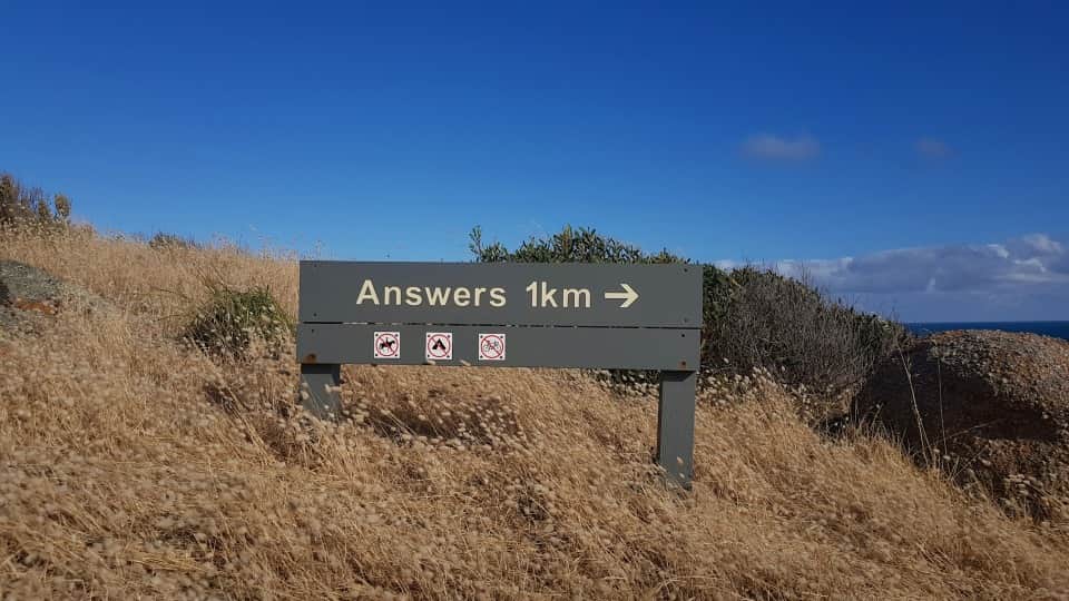 A sign in a park that says 'Answers 1km' with no entry signs for horses, camping and bicycles. Sign is planted within long dry grass blowing in the opposite direction to the sign