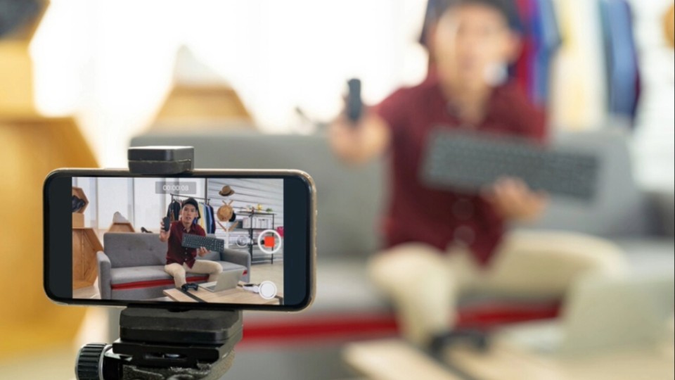 A tripod holding a phone horizontally recording a video of a man in the background sitting on a sofa.