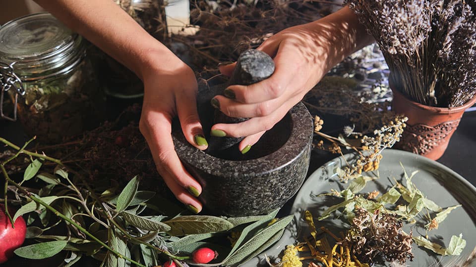 Hands grinding ingredients in a pestle and mortar with herbs and plants surrounding