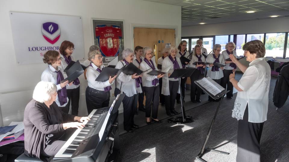 A group of people standing and singing together, one is sat playing a keyboard and one person is standing opposite the group, conducting them.