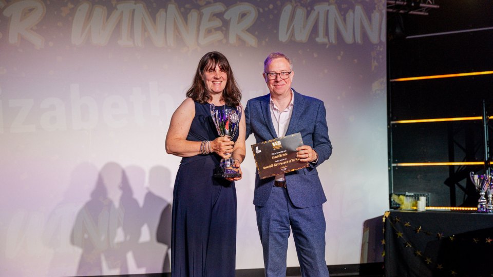 Two members of staff standing on stage at the Loughborough Experience Awards, one is holding a trophy and the other is presenting her with a certificate.