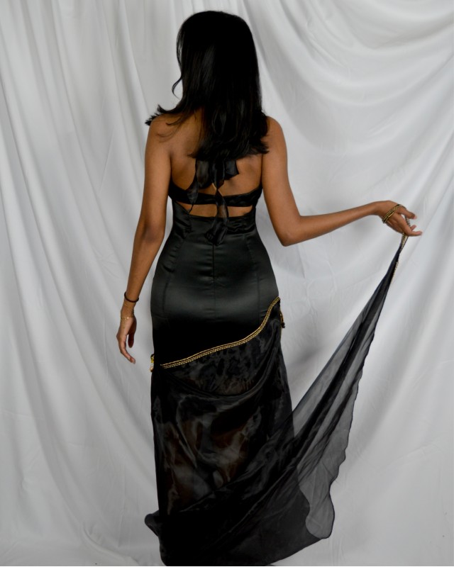 A woman wearing a long black dress standing with her back towards the camera in front of a white fabric backdrop.