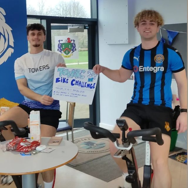 Two students on static bikes holding a sign reading 'Towers' 24-hour bike challenge'.