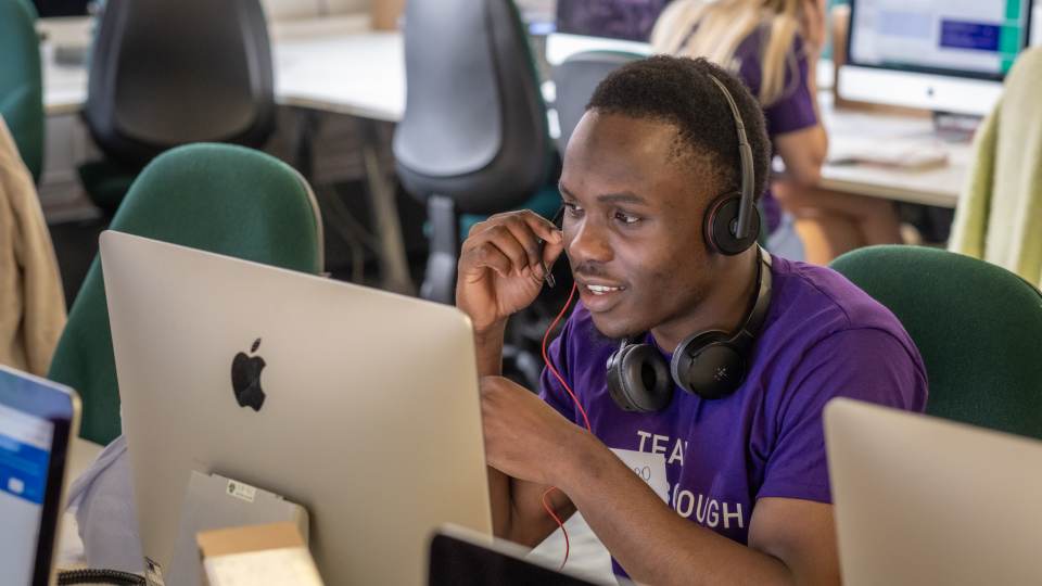 Photo of a person sat down in front of a computer wearing a headset mid-conversation