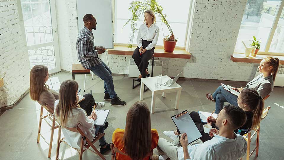 Two people having a discussion in a classroom setting with others sat around in a semicircle taking notes