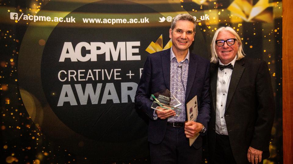 Two Loughborough Creative and Print Services team members holding a glass trophy standing in front of a black and gold awards backdrop reading 'ACPME CREATIVE + PRINT AWARDS'.