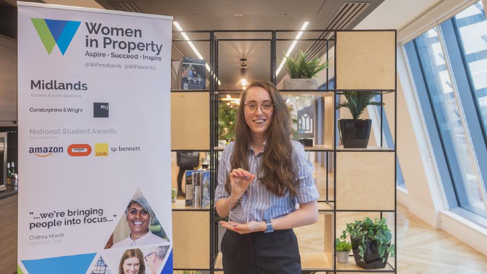 A student holding a glass trophy, standing next to a 'Women in Property' standing banner.