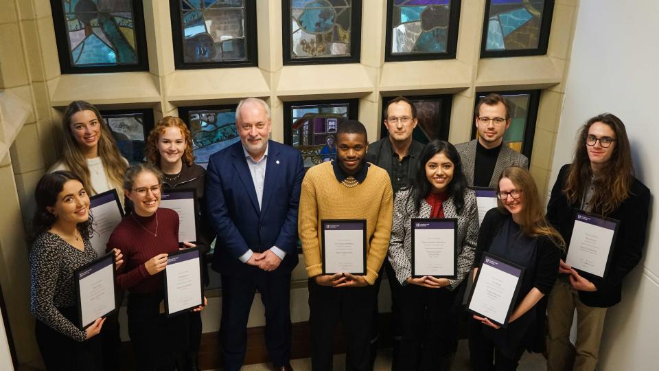 A group of students standing with Professor Nick Jennings in front of stained glass windows, holding framed certificates. 