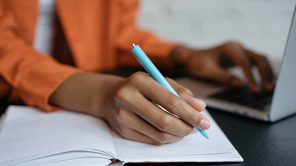 Close-up image of someone's hand with a pen writing notes in a book whilst using the other hand to type on a silver laptop