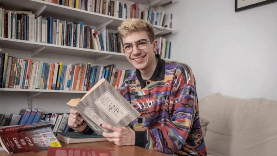 A student sitting in front of a bookshelf with a book open in their hands.