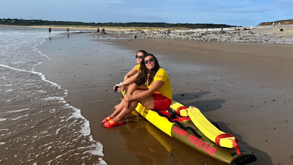 Beth Morgan smiling on a beach by the waterline in lifeguard gear