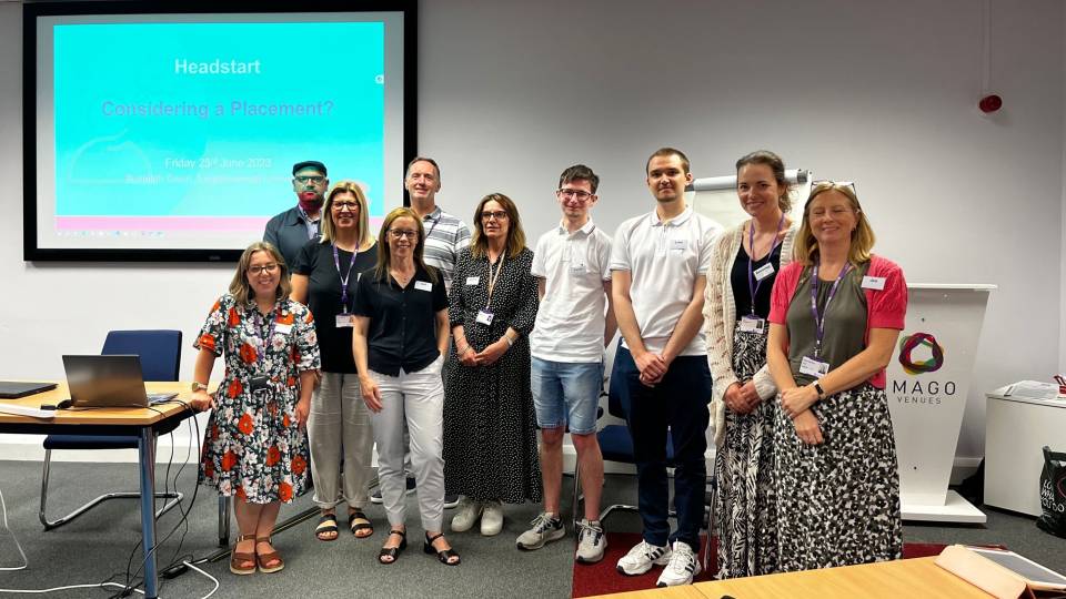 Photo of a group of staff at Loughborough University stood in a lecture/seminar room with a presentation screen behind them. They are all stood together smiling at the camera