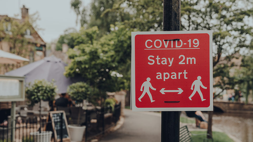 Photo of a red informative sign attached to a lamp post by a road with an icon of people staying 2m distance apart. 