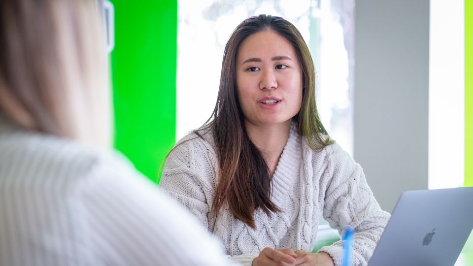 Photo of a female student sat down with a laptop talking to someone next to her