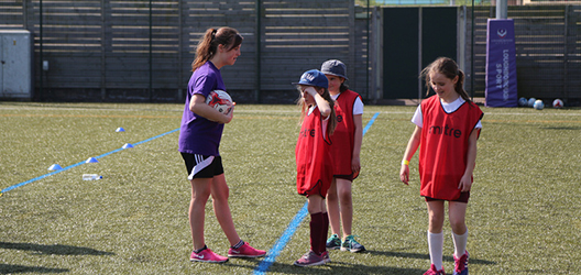 photo of a female student teaching football to some schoolchildren on a field