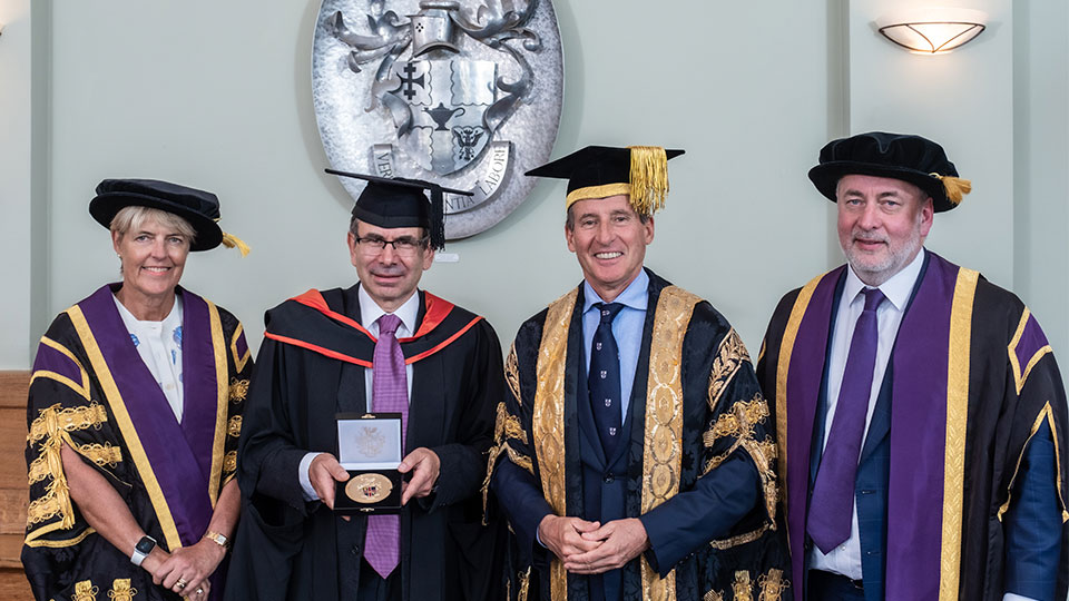 One woman and three men stand in academic robes. One of the men is holding a medal.