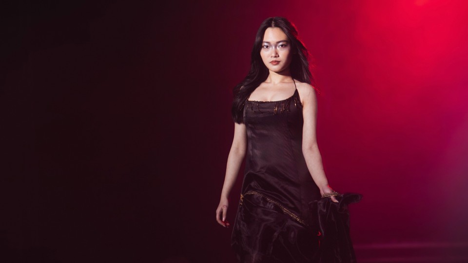 Model walking on a runway wearing a black dress with red lighting behind her.