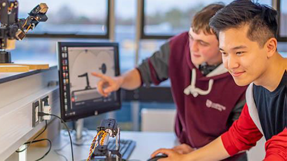 Photo of two students, one smiling, and the other pointing at a monitor with some other tech equipment surrounding them