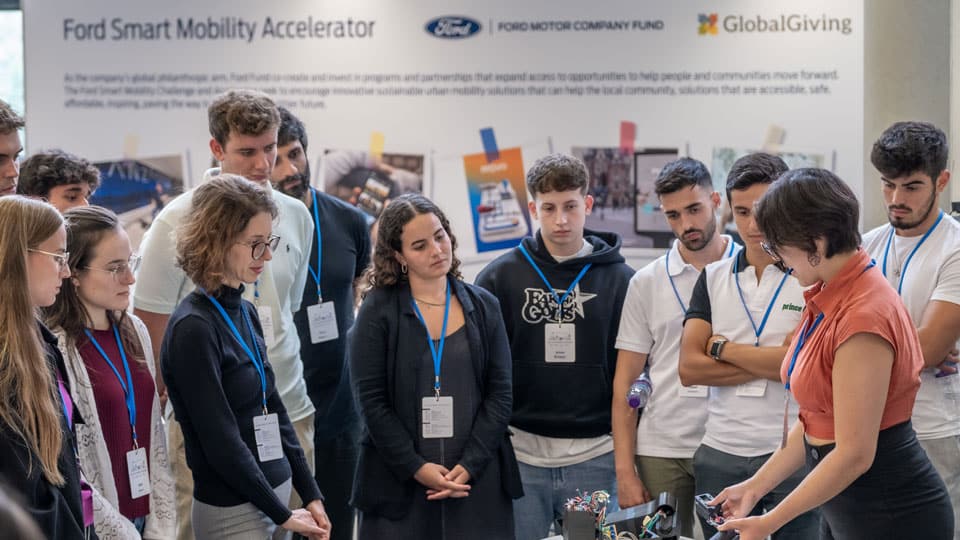 A group of people wearing blue lanyards watching a person doing a demonstration at the Ford Motor Company Fund Smart Mobility Accelerator event. 