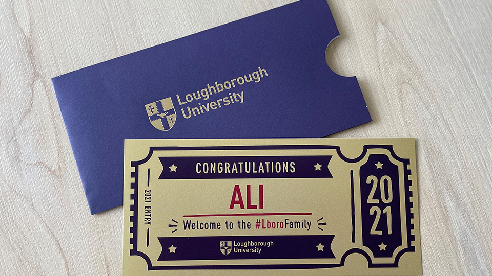 Photo of one of Loughborough's Golden Tickets accompanied by a purple envelope with the University logo, both laid on a table