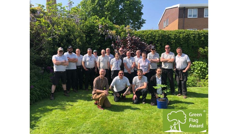 Loughborough Gardens team standing together on the grass with trees behind and a potted plant in front of them. The Green Flag Award logo is in the bottom right corner of the photo.